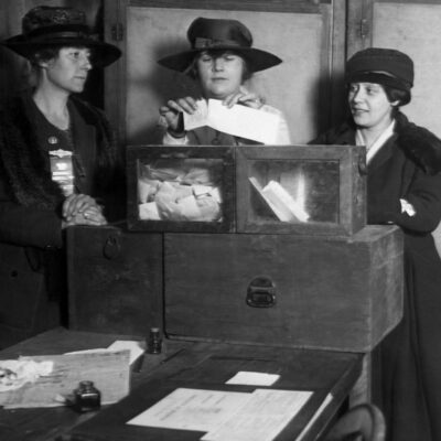 Three women count ballots beside a wooden ballot box in 1895