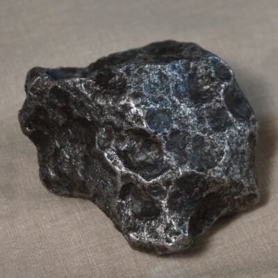 Dark metallic meteorite with rough, irregular surface on beige fabric