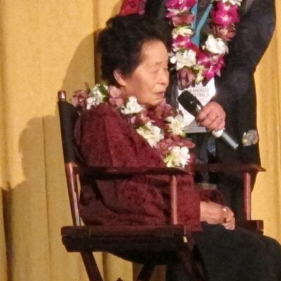 Keiko Fukuda seated and wearing flower leis during a public event