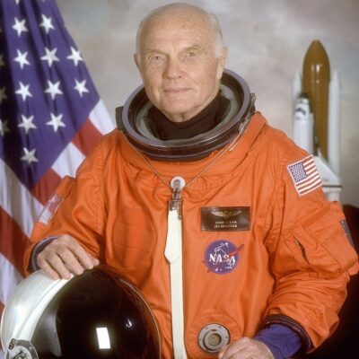 John Glenn in an orange NASA spacesuit holding a helmet, with an American flag and space shuttle in the background