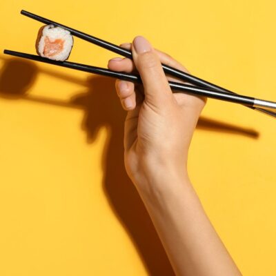 Hand holding chopsticks with a salmon sushi roll against yellow background