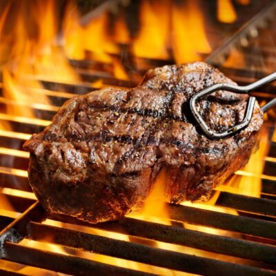 Seared steak cooking on a flaming grill with metal tongs