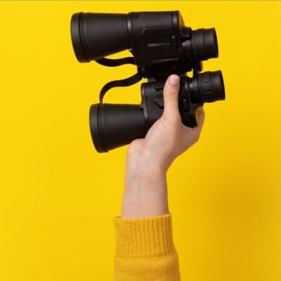 Hand holding black binoculars against a bright yellow background