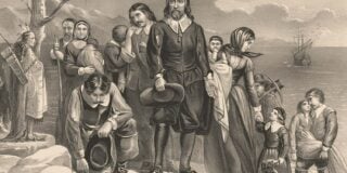 A group of Pilgrims, including families with children, standing on the shore with a Native American and the Mayflower in the background