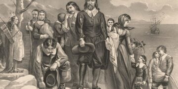 A group of Pilgrims, including families with children, standing on the shore with a Native American and the Mayflower in the background