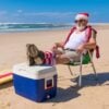 Man wearing a Santa hat and sunglasses sitting on a beach chair with feet on a cooler, surfboard nearby, ocean in the background