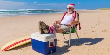 Man wearing a Santa hat and sunglasses sitting on a beach chair with feet on a cooler, surfboard nearby, ocean in the background