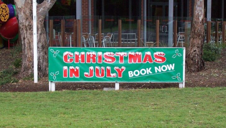 Sign promoting 'Christmas in July' celebration event with booking information, set in a grassy outdoor area near a building