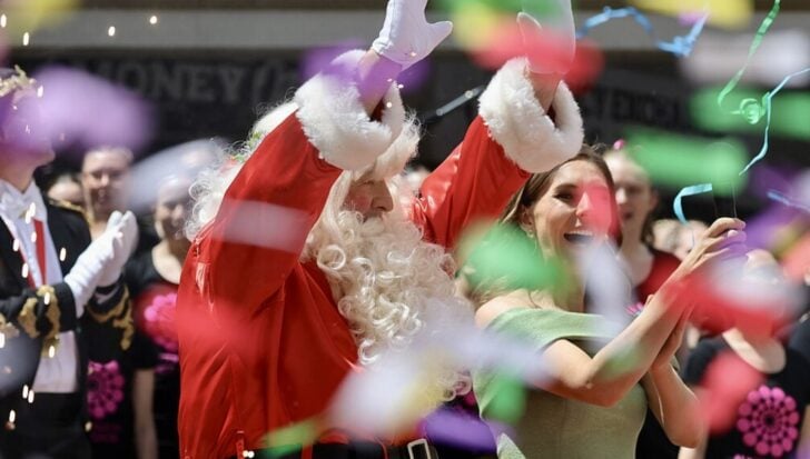 People celebrating outdoors with a man dressed as Santa Claus alongside a woman, surrounded by festive streamers and a crowd in the background