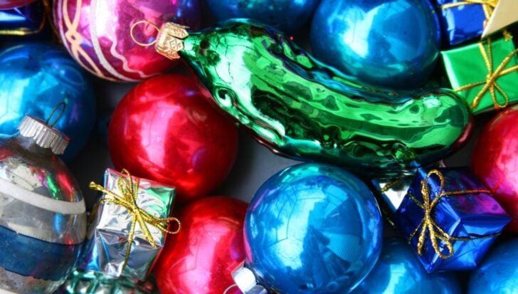 Green glass pickle ornament surrounded by colorful Christmas baubles and small wrapped gifts