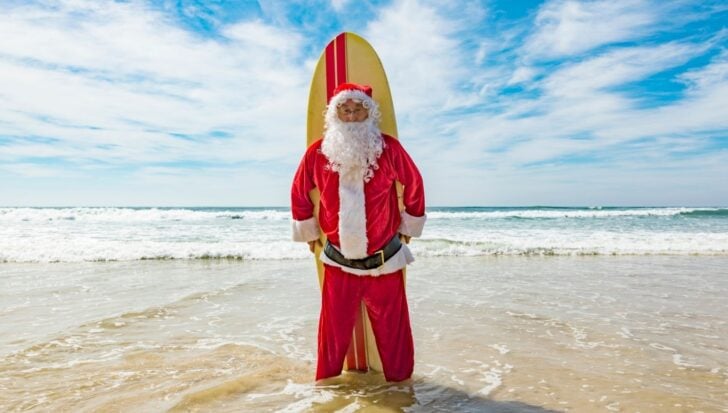 Santa Claus stands on a beach holding a surfboard with ocean waves in the background, depicting a unique summer celebration