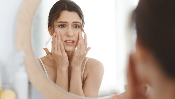 A person looks in the mirror, touching their face with a concerned expression