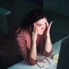 Person sitting in a dimly lit room, holding their head in their hands while looking at a computer screen, glasses and a pencil on the desk