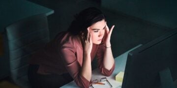 Person sitting in a dimly lit room, holding their head in their hands while looking at a computer screen, glasses and a pencil on the desk
