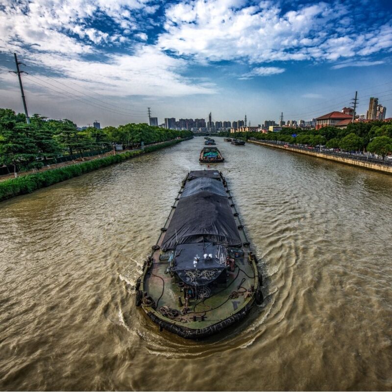 Cargo barges traveling along a wide canal lined with trees and buildings