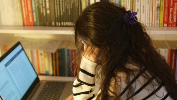 A woman with long hair and a striped sweater sits at a desk with her hand on her forehead, in front of a laptop and bookshelves