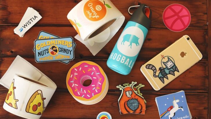 Various stickers displayed on a table, including designs of a donut, pizza slice, and a character in a space suit. A water bottle, phone with a sticker, and rolls of stickers are also visible.