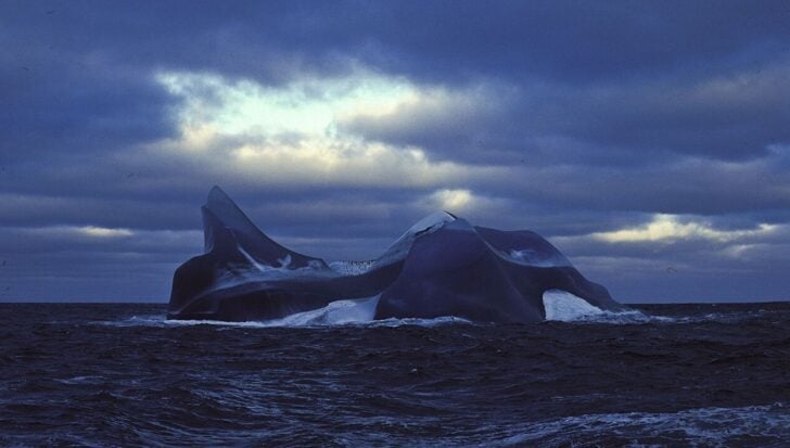 An iceberg with a dark hue floating in the Southern Ocean under a cloudy sky