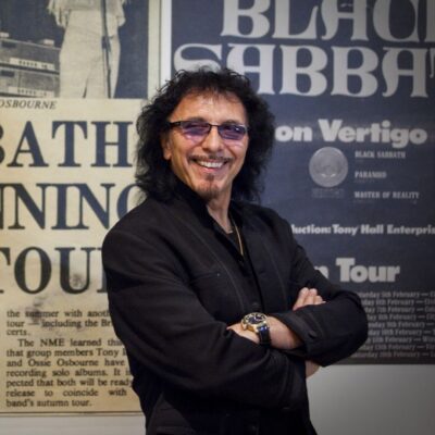 Tony Iommi smiling with folded arms in front of Black Sabbath posters