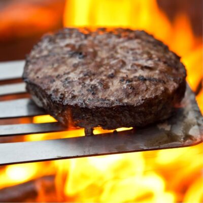 Grilled hamburger patty on a metal spatula over open flames