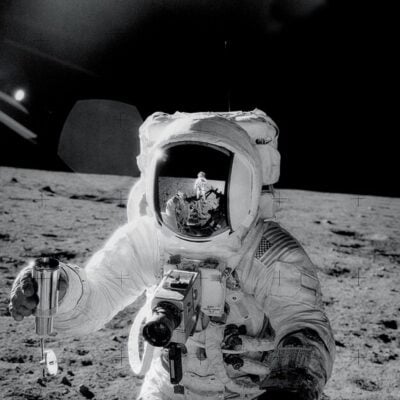 Astronaut on the Moon holding equipment with another astronaut reflected in helmet visor