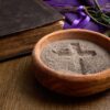 A wooden bowl with ashes forming a cross, an old bible, and a purple cloth