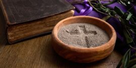 A wooden bowl with ashes forming a cross, an old bible, and a purple cloth