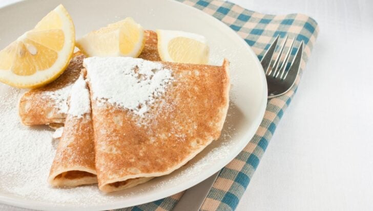 Plate of pancakes with powdered sugar and lemon wedges, placed on a checkered napkin with a fork