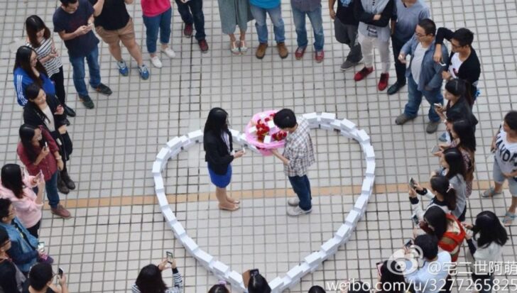 A man kneeling inside a heart made of 99 iPhone boxes, proposing to a woman holding a bouquet