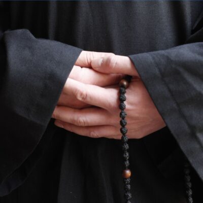 Hands folded in front of a black monastic robe, holding knotted prayer beads