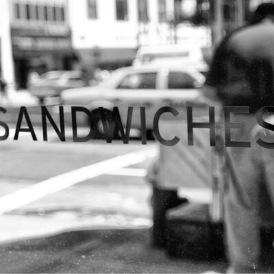 Storefront window with "sandwiches" written on it, reflecting a busy city street