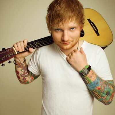 Ed Sheeran poses with an acoustic guitar slung over his shoulder, showing tattooed arms