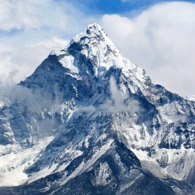Snow-covered, jagged mountain peak rises above clouds against a blue sky