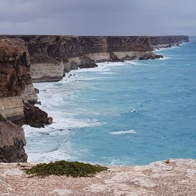 Rugged coastal cliffs overlook turquoise water and white waves under a cloudy sky