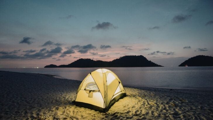 A glowing tent on a sandy beach at dusk, with an ocean view and distant silhouettes of islands