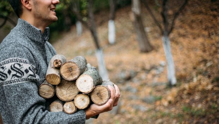Person carrying a stack of firewood in an outdoor setting with fall leaves in the background
