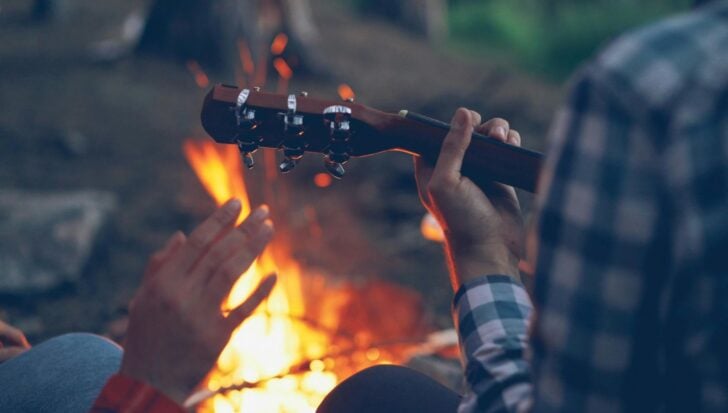 Two people sitting by a campfire, one playing a guitar