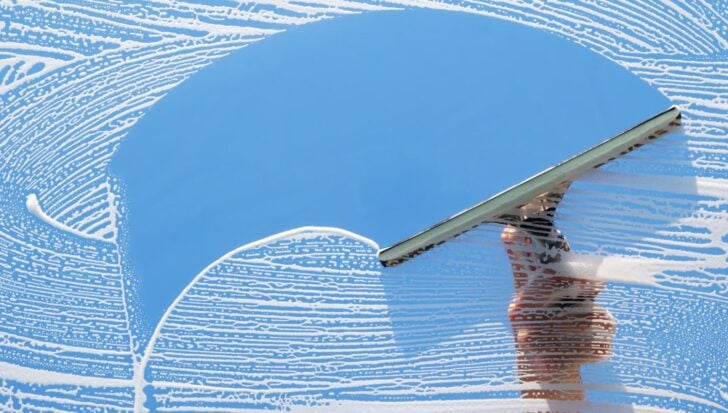 A hand operating a squeegee cleaning a glass surface with soap suds