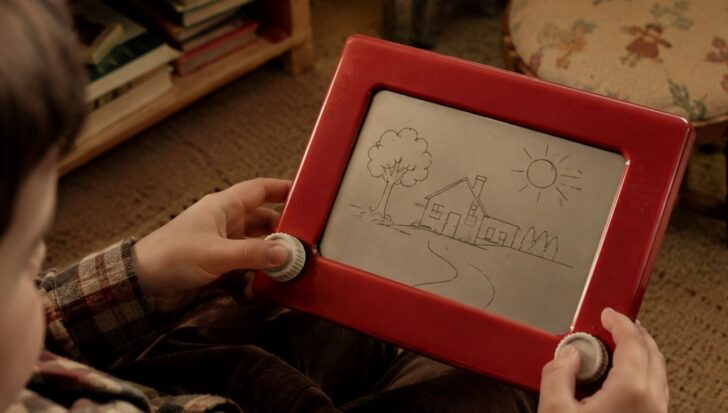 A person holds an Etch A Sketch displaying a drawing of a house, tree, and sun