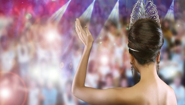 A woman with a tiara waves to a crowd in a colorful, spotlighted setting